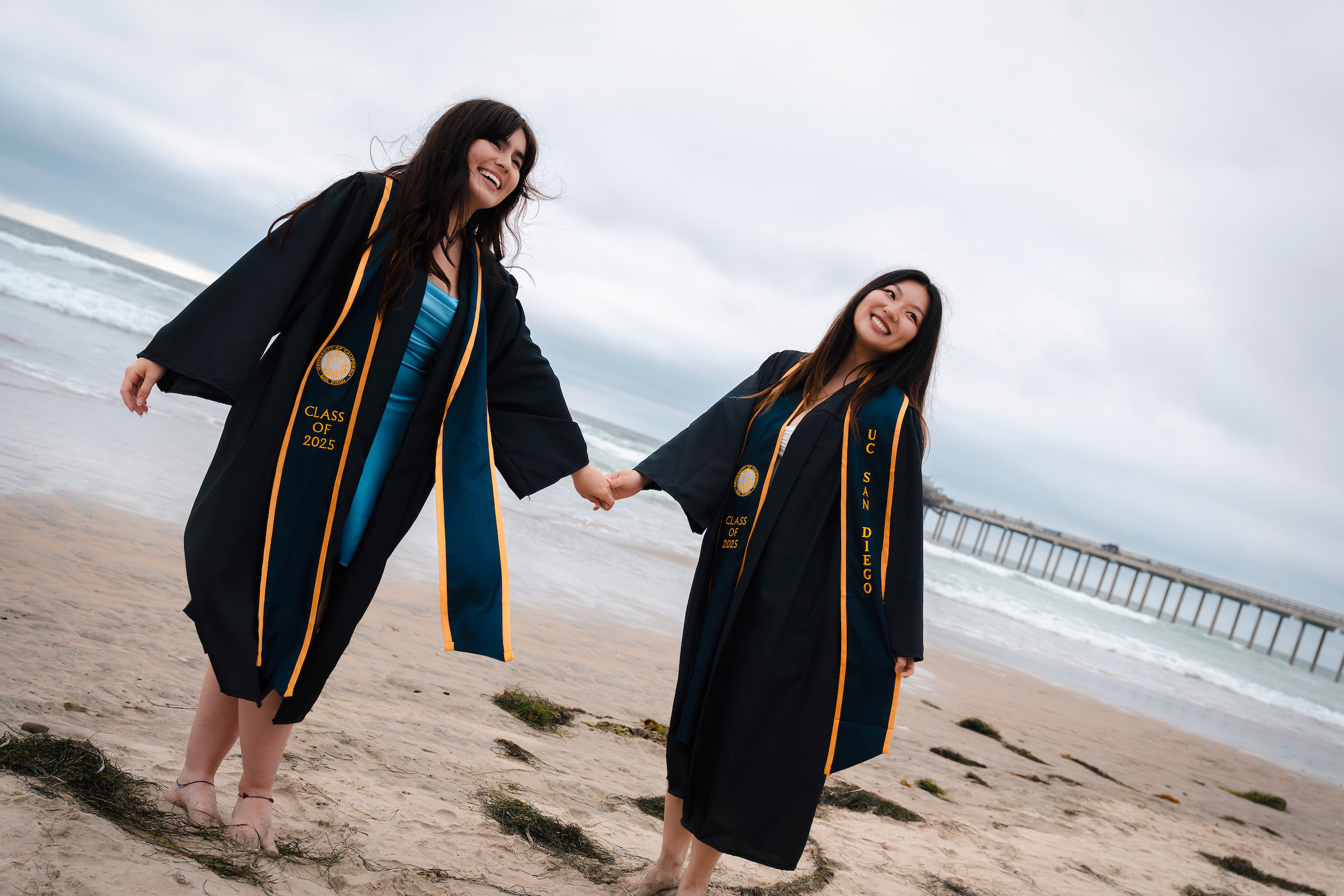 Two students in commencement regalia hold hands and smile on a beach, the ocean behind them.