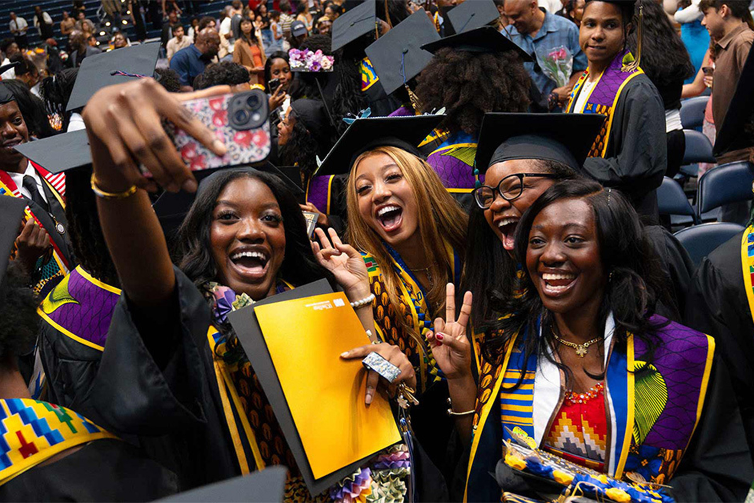 Four graduating students smile and cheer while taking a selfie in caps and gowns.