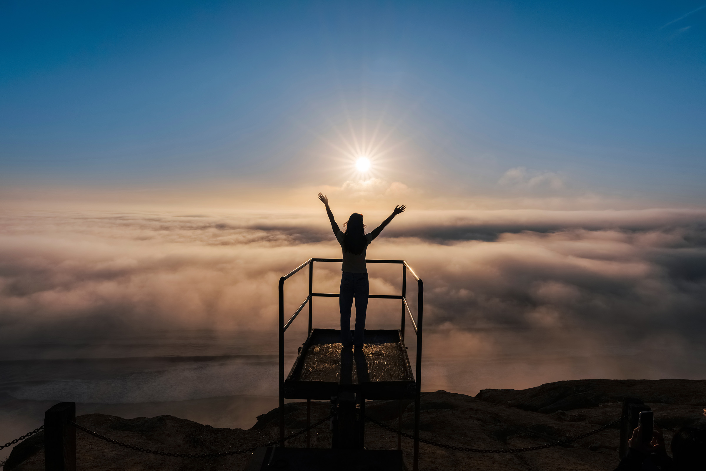 Person stands facing sun and clouds with hands reached to the sky