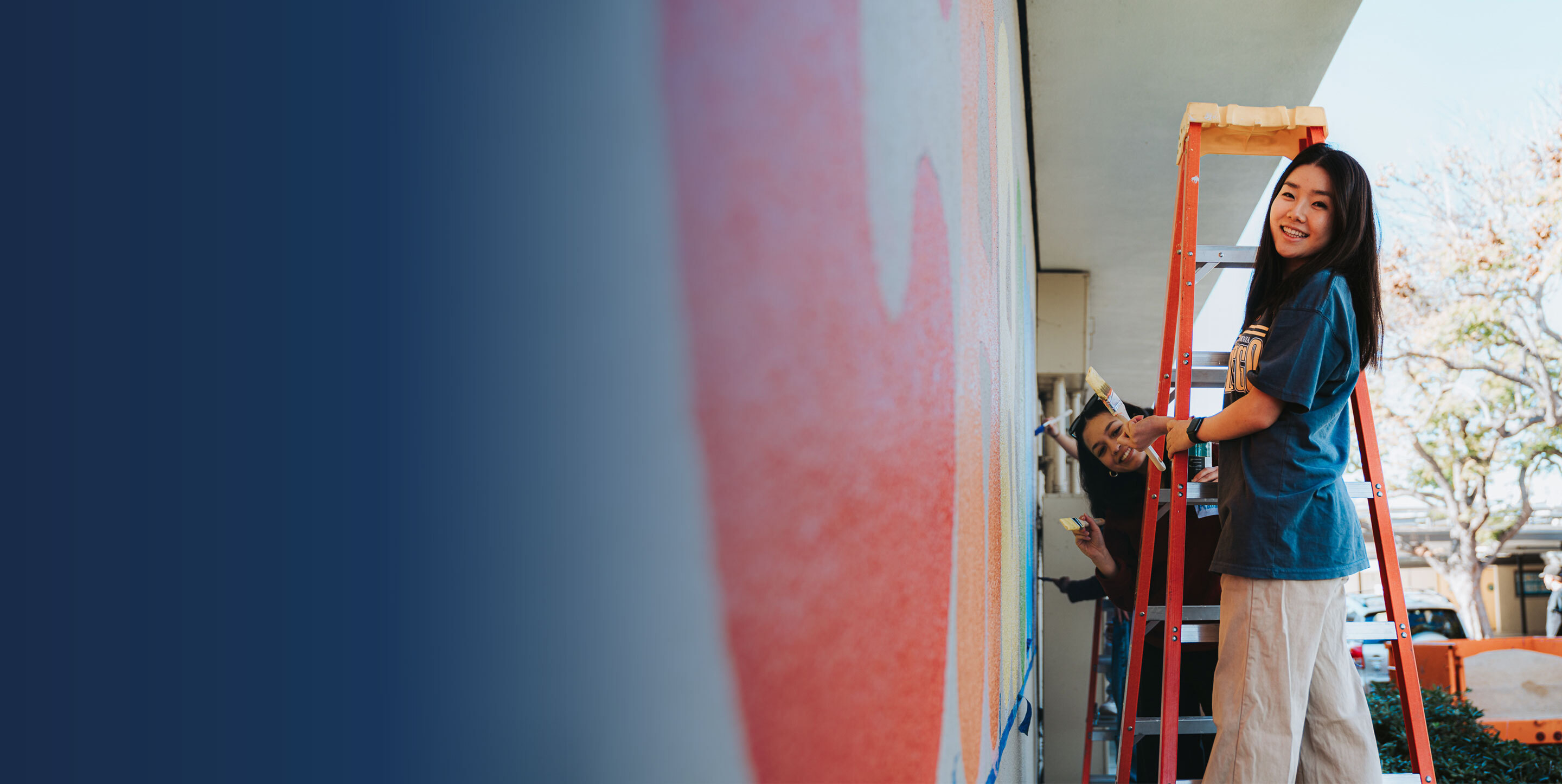 Students stand on ladder with paintbrushes next to wall mural